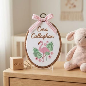 May include: An oval wooden embroidery hoop containing white fabric with the embroidered name Cora Callaghan, a pink flamingo, green palm leaves, and small pink flowers. A pink and white gingham ribbon bow is tied at the top of the hoop.