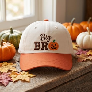 May include: A cream-colored baseball cap with an orange brim. The front features the brown embroidered text "Big BRO" alongside a small orange jack-o'-lantern pumpkin graphic with a green stem, centered on the crown of the hat.