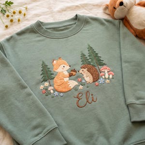 May include: A sage green crewneck sweatshirt featuring an embroidered woodland scene with a fox holding an acorn, a hedgehog, pine trees, mushrooms, and small flowers. The name Eli is embroidered in brown cursive script below the animals.