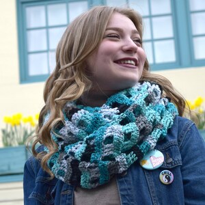 May include: A woman wearing a blue denim jacket with a teal, gray, and white crocheted infinity scarf. She is wearing two pins on her jacket, one with the words "Heavy Eats" and the other with the word "Chill".