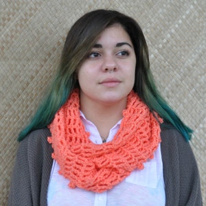 May include: A woman with teal hair is wearing a coral crochet infinity scarf. The scarf is made with a delicate open stitch pattern.