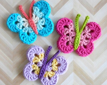 May include: Three crocheted butterfly appliques in shades of blue, pink, and purple. Each butterfly has a different color scheme, with details like the antennae and center of the wings in contrasting colors. The butterflies are handmade.