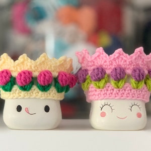 May include: Two white ceramic mugs with smiling faces, each wearing a crocheted crown. One crown is yellow with pink flowers, the other is pink with purple flowers.