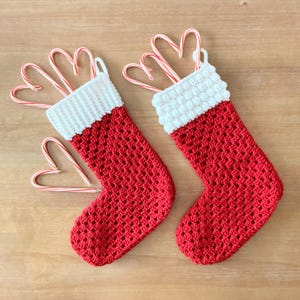 May include: Two handmade crochet Christmas stockings in a vibrant red yarn with white cuffs. The left stocking features a ribbed white cuff, while the right stocking has a textured bobble stitch cuff. Each stocking is accented with candy canes.