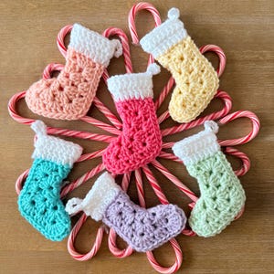May include: Six small crocheted Christmas stockings in pastel shades of peach, yellow, bright pink, turquoise, lavender, and light green, each featuring a white cuff, arranged in a circular pattern over a star-shaped cluster of red and white candy canes.