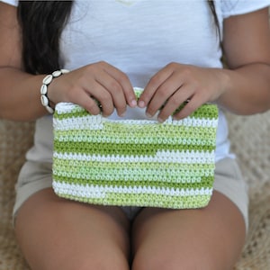 May include: A green and white striped crocheted handbag with a handle. The bag is being held by a person.