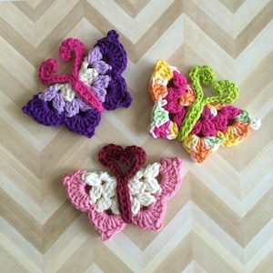 May include: Three colourful, crocheted butterfly appliques. One is purple, lavender, and pink. Another is pink, orange, yellow, and green. The third is pink and white. The butterflies are handmade and sit on a light brown, chevron-patterned surface.