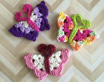 Puede incluir: Tres apliques de mariposa de ganchillo coloridos. Uno es morado, lavanda y rosa. Otro es rosa, naranja, amarillo y verde. El tercero es rosa y blanco. Las mariposas están hechas a mano y se encuentran sobre una superficie marrón claro con estampado de chevrón.