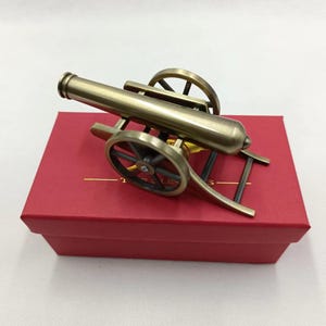 May include: A miniature bronze cannon with black wheels and a detailed barrel, resting on a red box. The cannon is a historical replica, showcasing a classic design. The overall aesthetic is vintage and decorative.