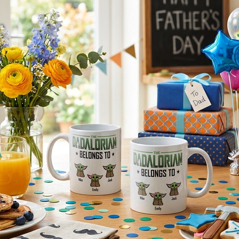 May include: Two white ceramic mugs featuring the text THE DADALORIAN BELONGS TO and illustrations of three small green creatures labeled Mike, Emily, and Jack. The mugs are positioned on a table with colorful confetti, a glass of orange juice, and gifts.