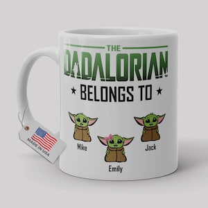 May include: A white ceramic mug featuring the green text THE DADALORIAN BELONGS TO above three cartoon green creatures in brown robes. The names Mike, Emily, and Jack are printed below each character. A small tag with a US flag is attached.