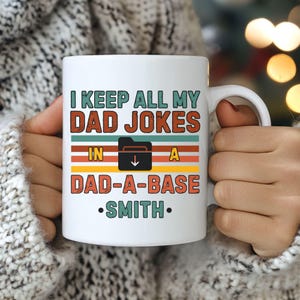 Könnte beinhalten: Weiße Keramik-Tasse mit dem Text "I KEEP ALL MY DAD JOKES IN A DAD-A-BASE SMITH" in einem Retro-Design. Die Tasse hat einen weißen Henkel und wird von einer Person gehalten, die einen weißen Pullover trägt.