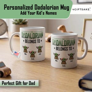 May include: Two white ceramic mugs featuring the text THE DADALORIAN BELONGS TO in green and black lettering. Below the text are three cartoon illustrations of green creatures with large ears, labeled with the names Mike, Emily, and Jack.