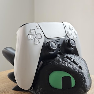 Może przedstawiać: Czarny i zielony stojak na kontroler z motywem smoka z białym i czarnym kontrolerem PlayStation 5 na górze. Stojak przedstawia głowę smoka z zielonymi oczami. Kontroler ma symetryczny design.