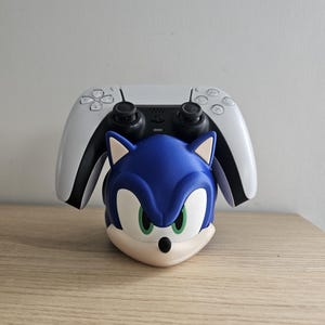 Op de afbeelding: Een blauw-witte Sonic the Hedgehog controllerhouder met een witte PlayStation 5 controller erop. De houder toont Sonic's gezicht met groene ogen en een beige snuit. De controller is wit met zwarte details.