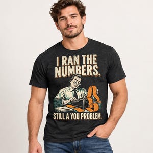 May include: A charcoal grey short-sleeve t-shirt featuring a graphic of a person using an adding machine with a long paper tape. The text reads I RAN THE NUMBERS. STILL A YOU PROBLEM. The design uses a vintage colour palette of tan and black.