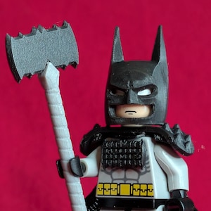 Op de afbeelding: Een Lego Batman-figuur met een zwarte kap, cape en harnas, die een grijze bijl vasthoudt. De figuur heeft een witte torso met een geel en zwart riemontwerp. De achtergrond is effen rood.