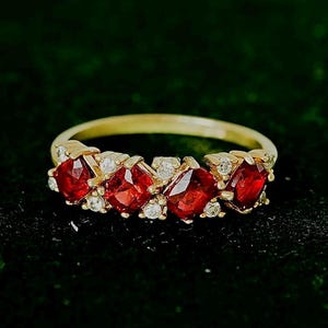 May include: A gold ring with five vivid red gemstones, set between smaller white stones. The ring's band is polished gold, contrasting with the deep red of the gems. The ring is displayed against a dark background, highlighting the jewellery.