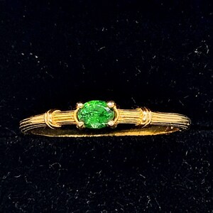 Könnte beinhalten: Ein goldener Ring mit einem leuchtend grünen, ovalen Edelstein. Der Ring hat ein zartes Band mit gerippten Details und eine Vier-Zacken-Fassung. Der Ring ist vor einem dunklen Hintergrund platziert.