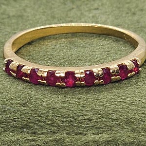 Peut inclure: Une bague en or ornée d'une rangée de pierres précieuses rouges vives. La monture dorée entoure le doigt, et les pierres rouges sont disposées sur le dessus. La bague est présentée sur un fond vert.