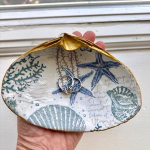 May include: A decorative seashell ring dish with a gold-painted rim. The interior features blue starfish, coral, and seashell designs on a white background. Two silver rings rest inside the dish.