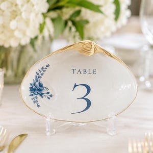 May include: A white seashell table number card holder with a gold-painted rim. The shell features a blue floral illustration on the left side and the text "TABLE 3" printed in dark blue ink in the center, supported by a clear acrylic stand.