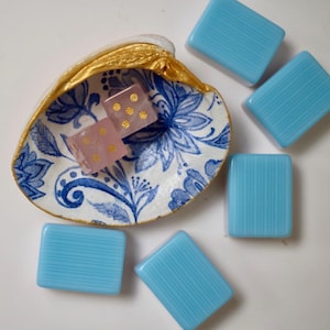 May include: A decorative seashell dish with a blue and white floral pattern and gold-painted edges, containing two translucent pink dice with gold pips. Five rectangular light blue tiles with vertical ribbed textures are arranged around the shell.