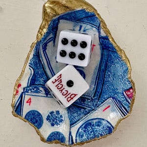 May include: A decorative oyster shell dish with a gold-painted rim, holding two white dice. The dish features a blue and white playing card design. One die shows six dots, the other shows one dot and the word "Bicycle".
