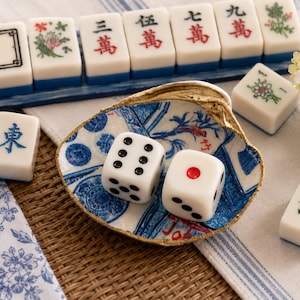 May include: A decorative seashell dish with a gold-painted rim and blue patterned interior, containing two white six-sided dice. Surrounding the dish are several white Mahjong tiles with black, red, blue, and green characters and floral designs.