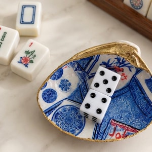 May include: A decorative clam shell dish with a gold-painted rim and a blue and white patterned interior. Inside the shell are two white six-sided dice with black pips. Nearby are three white mahjong tiles with green, red, and blue markings.