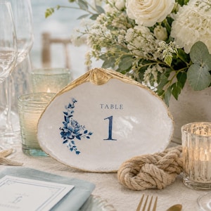 May include: A white seashell table number holder with a gold rim and blue floral design. The number "1" is printed in blue. A rope knot and gold cutlery are also visible.