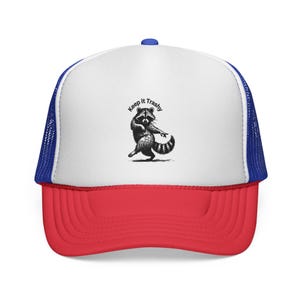May include: A trucker hat with a white front panel, a red bill, and a blue mesh back. The hat features a black and white illustration of a raccoon with the text "Keep it Trashy" above the raccoon.
