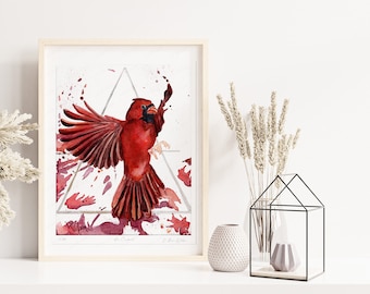 May include: A framed watercolor print of a vibrant red cardinal bird with outstretched wings. The artwork features a geometric design and splashes of red and maroon, set against a white background. The print is displayed on a white shelf.