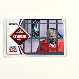 May include: A novelty card featuring an image of a person in an orange jumpsuit behind bars. The card has the text "RESIDENT ANNUAL AMERICA 250" and "Official White House Photo by Daniel Terok". The card is encased in a clear plastic sleeve.