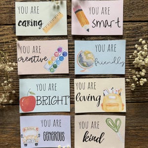 May include: Eight colorful cards with encouraging messages and illustrations, including a ruler, pencil, paint palette, globe, apple, backpack, school bus, and scissors. The cards read "You are caring, smart, creative, friendly, bright, loving, generous, kind."