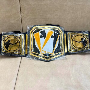 May include: A black and gold championship belt with a cornhole board design. The belt has a black strap with gold accents and two side plates with a silhouette of a person throwing a bean bag. The center plate features a cornhole board with the text "Cornhole Champion".