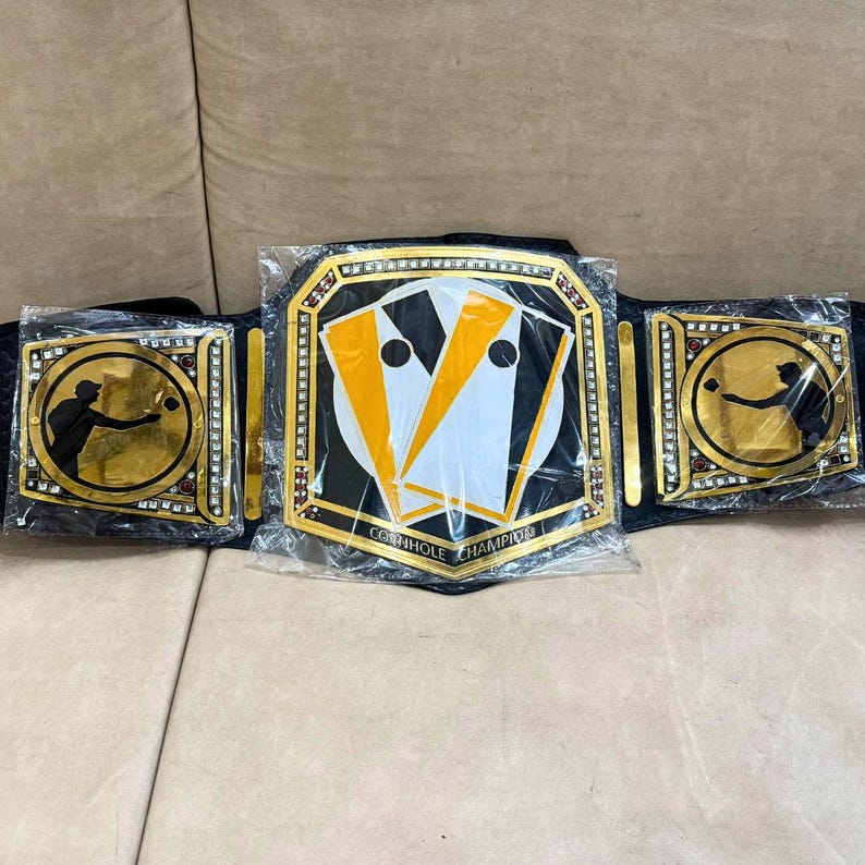 May include: A black and gold championship belt with a cornhole board design. The belt has a black strap with gold accents and two side plates with a silhouette of a person throwing a bean bag. The center plate features a cornhole board with the text "Cornhole Champion".