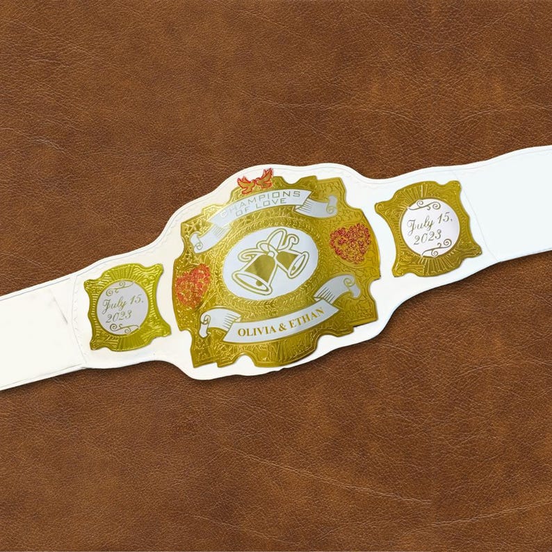 May include: White wedding championship belt with gold accents and the words "Champions of Love" and "Olivia & Ethan". The belt also has two gold medallions with the date "July 15, 2023".