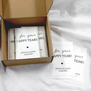 May include: A brown cardboard box filled with white packets. Each packet is printed with the words "for your HAPPY TEARS", the names "JESSICA & JASON" and the date "12.06.2025" in black. A small black heart is also printed on the packets.
