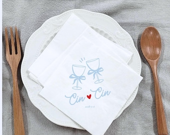 May include: White paper napkins with a light blue graphic of two wine glasses clinking, tied with bows. The napkins are on a white plate with a wooden fork and spoon on either side. The text "Cin Cin" and a red heart are printed on the napkins.