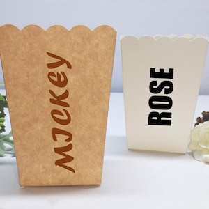 Peut inclure: Deux boîtes à pop-corn aux bords festonnés. L'une est marron avec le nom "MJekey" en écriture marron. L'autre est blanche avec le nom "ROSE" en lettres noires. Une fleur blanche et de la verdure sont en arrière-plan.