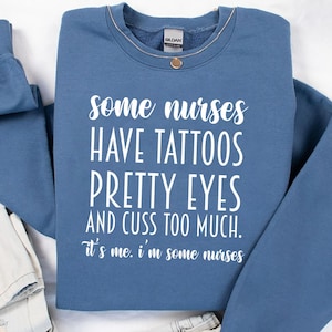 May include: Indigo blue sweatshirt with the text "some nurses HAVE TATTOOS PRETTY EYES AND CUSS TOO MUCH. it's me. i'm some nurses" in white. The sweatshirt is folded with a pair of light-wash jeans.