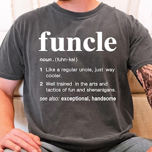 May include: Dark gray t-shirt with the word "funcle" in large white letters. The shirt includes a definition of "funcle" as a cooler uncle, with additional text describing the term. The shirt is a casual style.