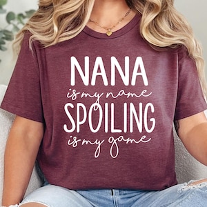 May include: Burgundy t-shirt with the text "NANA is my name, SPOILING is my game" in white. The shirt has a crew neck and short sleeves. The person is wearing blue jeans.