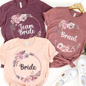 Puede incluir: Tres camisetas en tonos rosa y burdeos, con dise&ntilde;os de coronas florales. Las camisetas muestran las palabras "Team Bride", "Braut" y "Bride" en escritura blanca. Zapatillas blancas son visibles en la imagen.