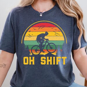 May include: Dark blue t-shirt featuring a cyclist silhouetted against a sunset design with the text "OH SHIFT" in yellow. The graphic includes a mountain bike, birds, and trees, creating a nature-themed design.