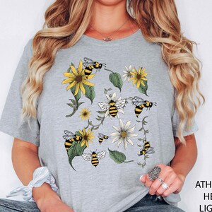 May include: Light grey t-shirt featuring a floral and bee design. The graphic includes yellow sunflowers, white daisies, green leaves, and black and yellow bees. The text "ATHLETHICH HEATHER LIGHT GREY" is printed on the right side of the image.