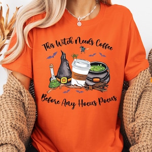 May include: Orange t-shirt with a Halloween-themed graphic. The design includes a witch's hat, a coffee cup, a cauldron, and the text "This Witch Needs Coffee Before Any Hocus Pocus". Bats and a witch flying on a broom are also featured.