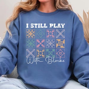 May include: A blue crewneck sweatshirt featuring a graphic print with the text I STILL PLAY With Blocks and a grid of twelve colorful, distressed-style quilt block patterns in shades of yellow, green, pink, and blue on the front center.