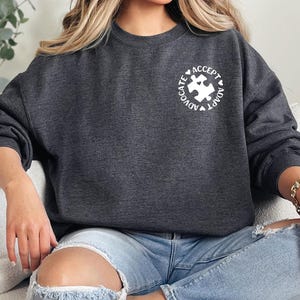 May include: Dark gray sweatshirt with a white puzzle piece graphic inside a circle with the words "Advocate, Accept, Adapt". The sweatshirt is a crew neck style and is made of a soft material.